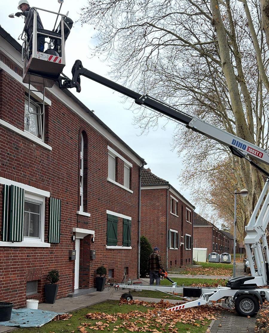 Anhänger-Arbeitsbühne Dino 160T11 bei Dacharbeiten an rotem Backsteinhaus in Wohnstraße, ausgefahrener Teleskoparm mit Arbeitskorb, herbstliche Straßenszene mit Laub.
