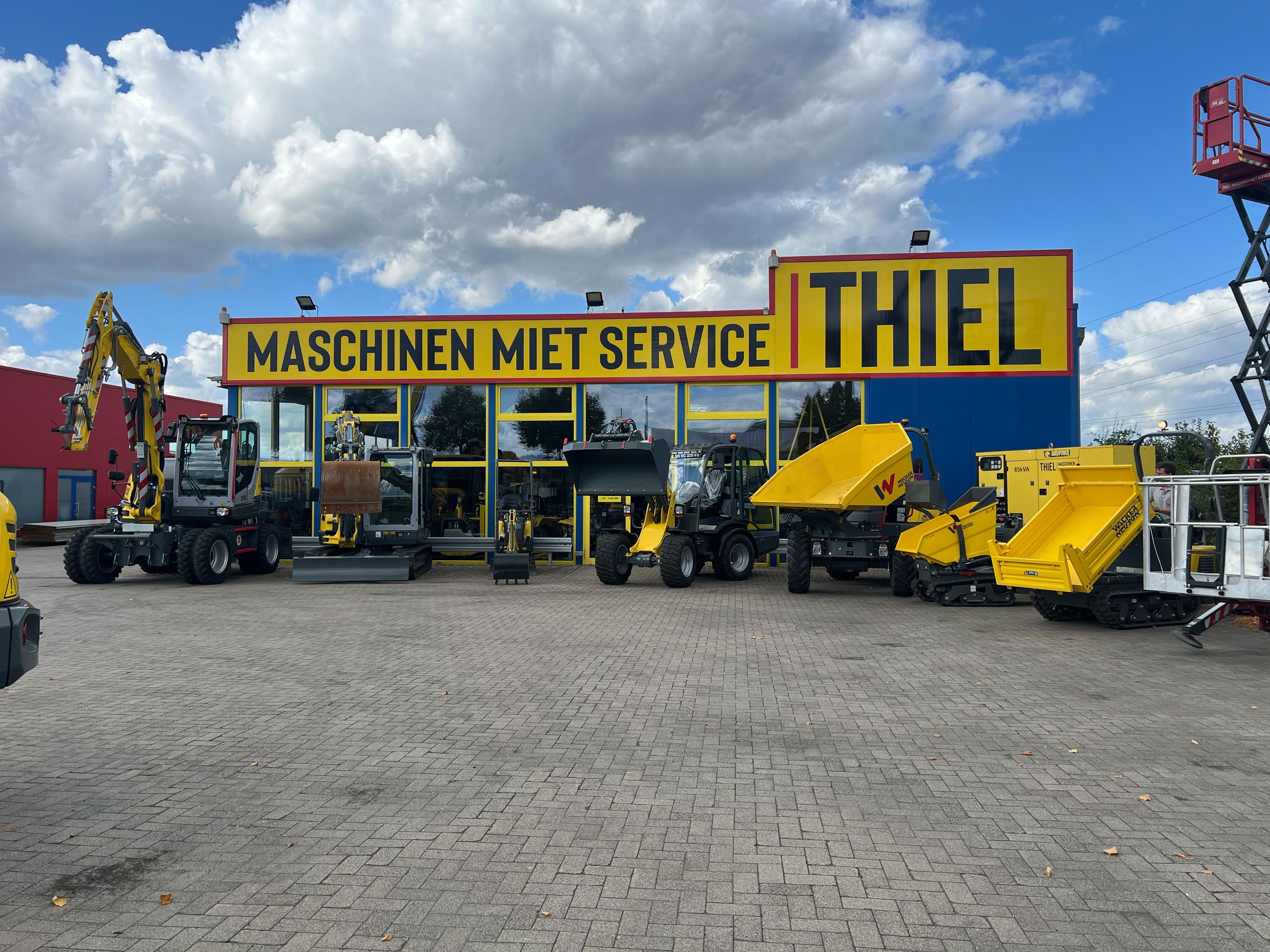 Baumaschinenverleih Ithiel mit blauem Gebäude und gelbem Schild, gelbe Baumaschinen wie Bagger und Asphaltfertiger auf gepflastertem Hof, bewölkter Himmel
