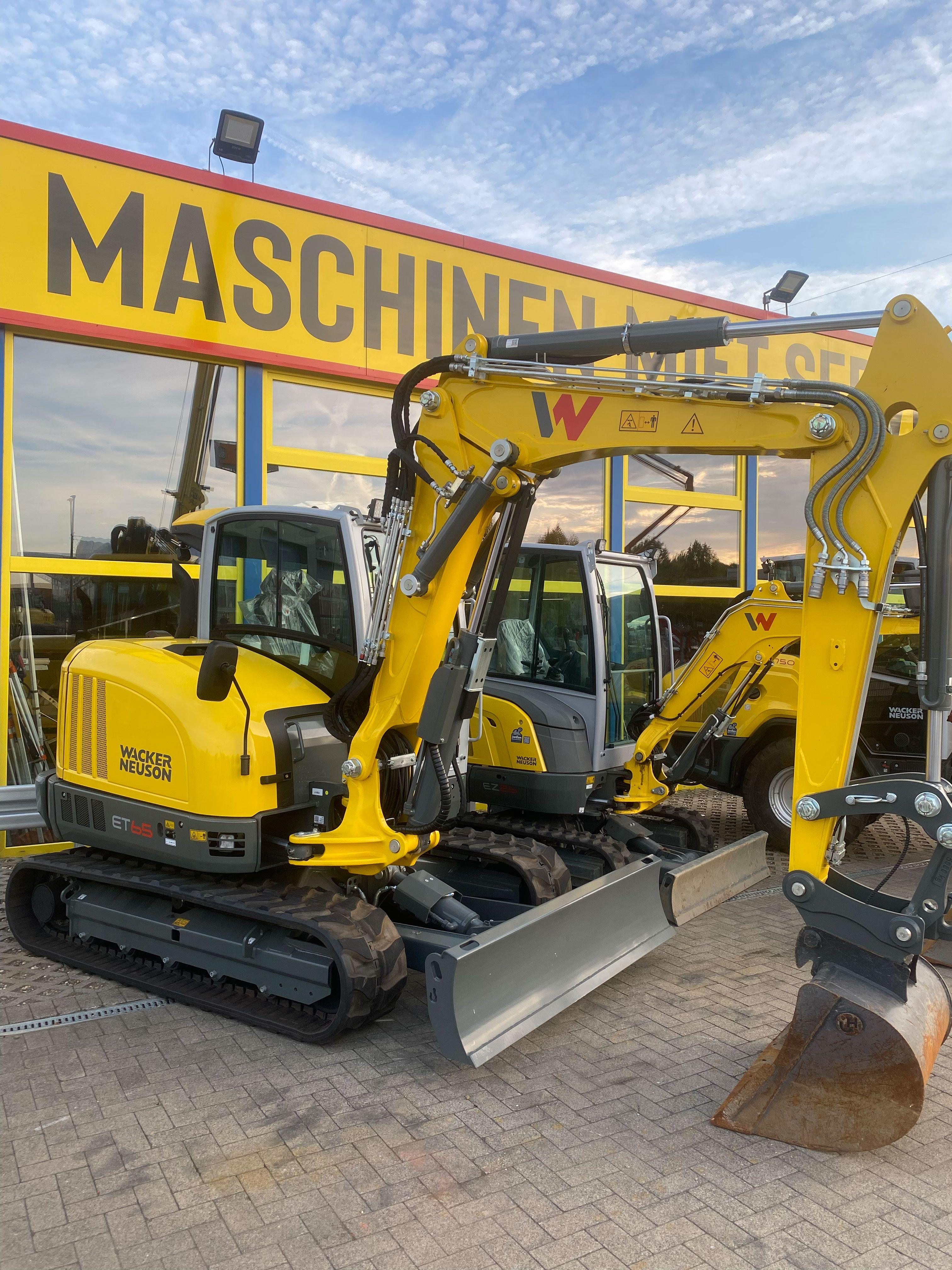Gelber Wacker Neuson Minibagger ET45 mit ausgefahrenem Baggerarm und Schaufel vor Maschinenverleih-Gebäude, weitere gelbe Baufahrzeuge im Hintergrund sichtbar.