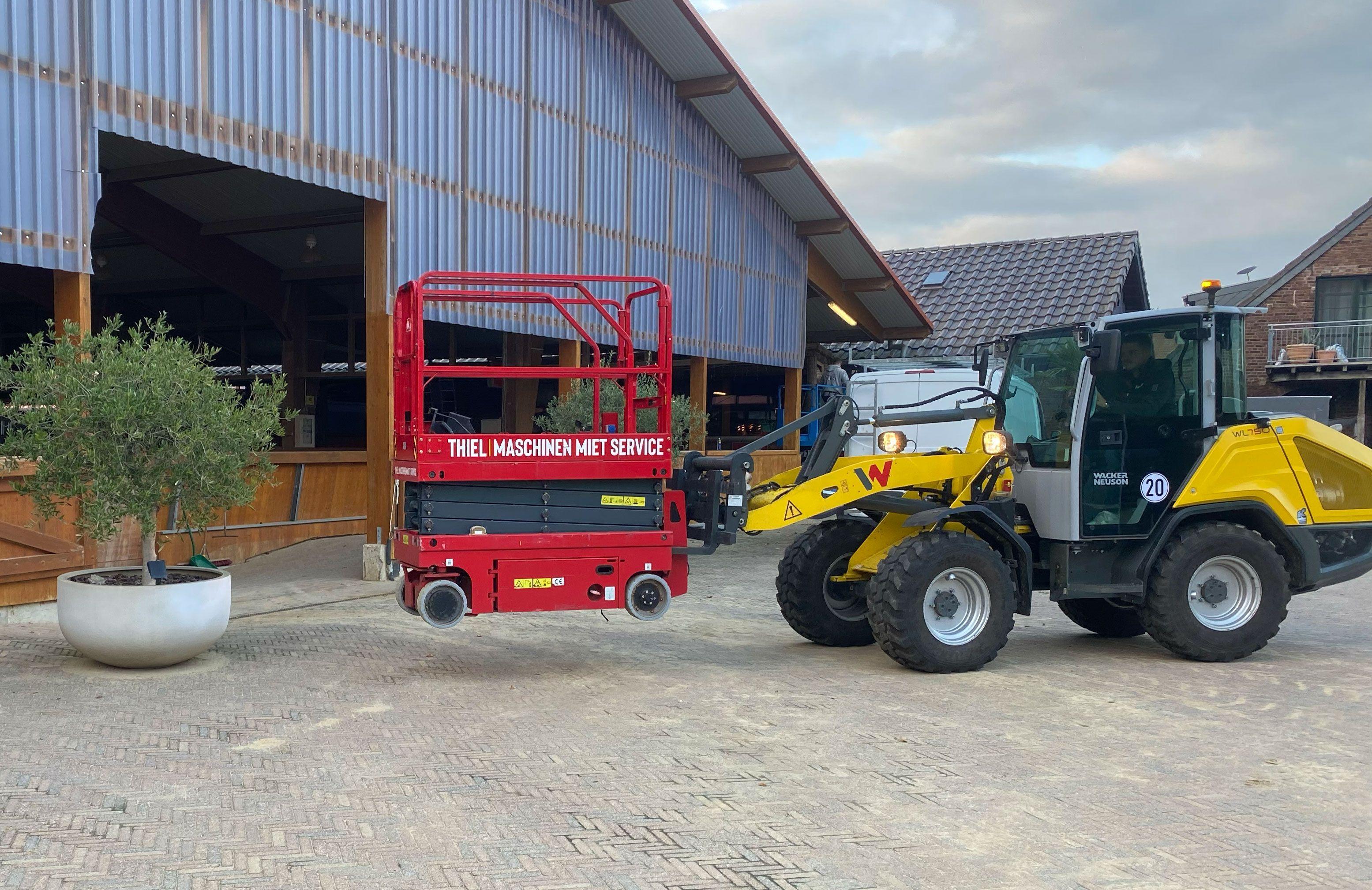 Rote Scherenarbeitsbühne von Thiel Maschinen Miet Service und gelber Wacker Neuson Radlader vor moderner Lagerhalle mit Wellblechfassade.