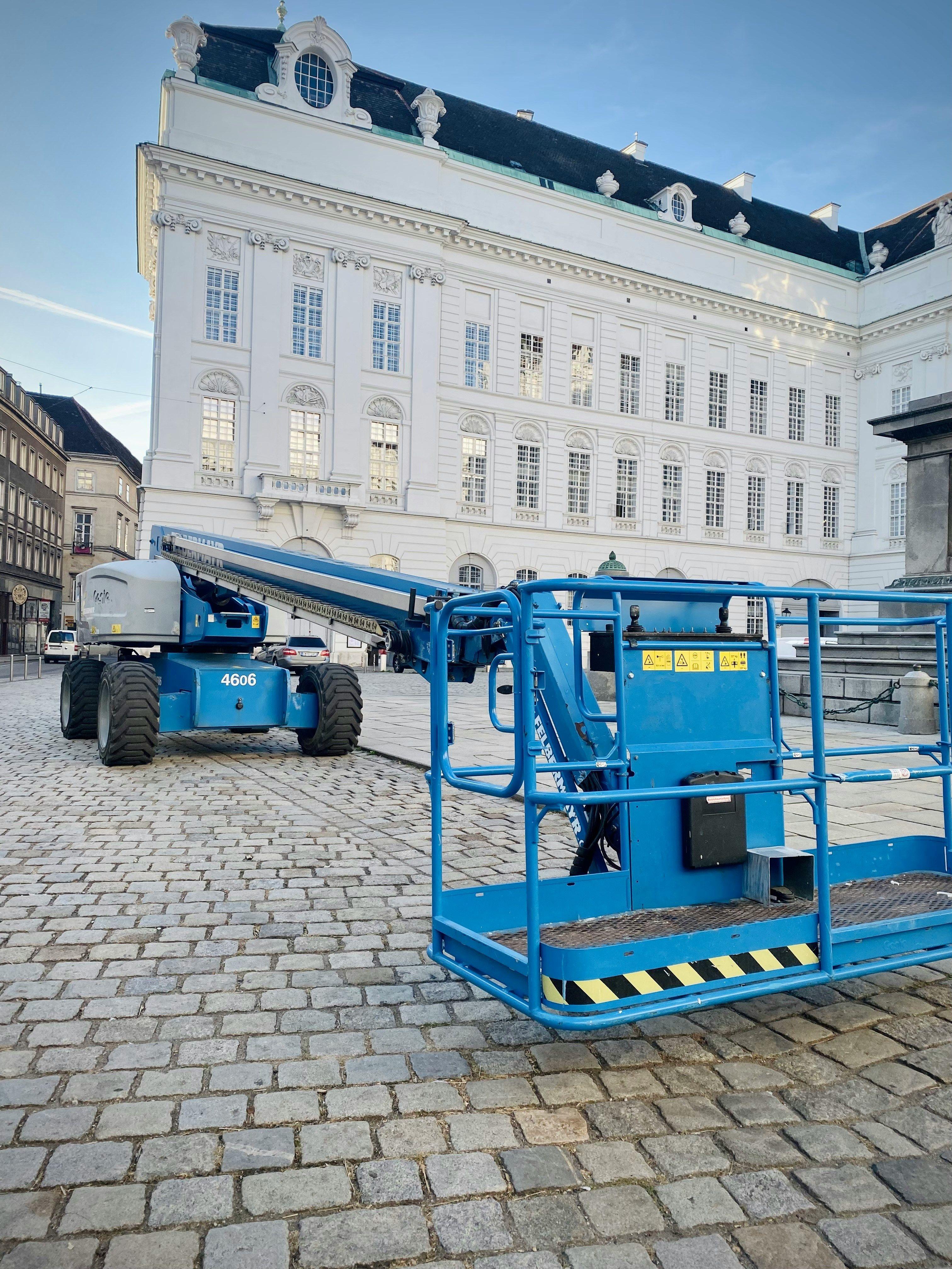 Blaue Gelenkteleskopbühne und Scherenhebebühne stehen auf einem historischen Kopfsteinpflasterplatz vor einem prächtigen weißen Barockpalast mit dunklem Schieferdach und ornamentalen Fassadenelementen unter blauem Himmel.