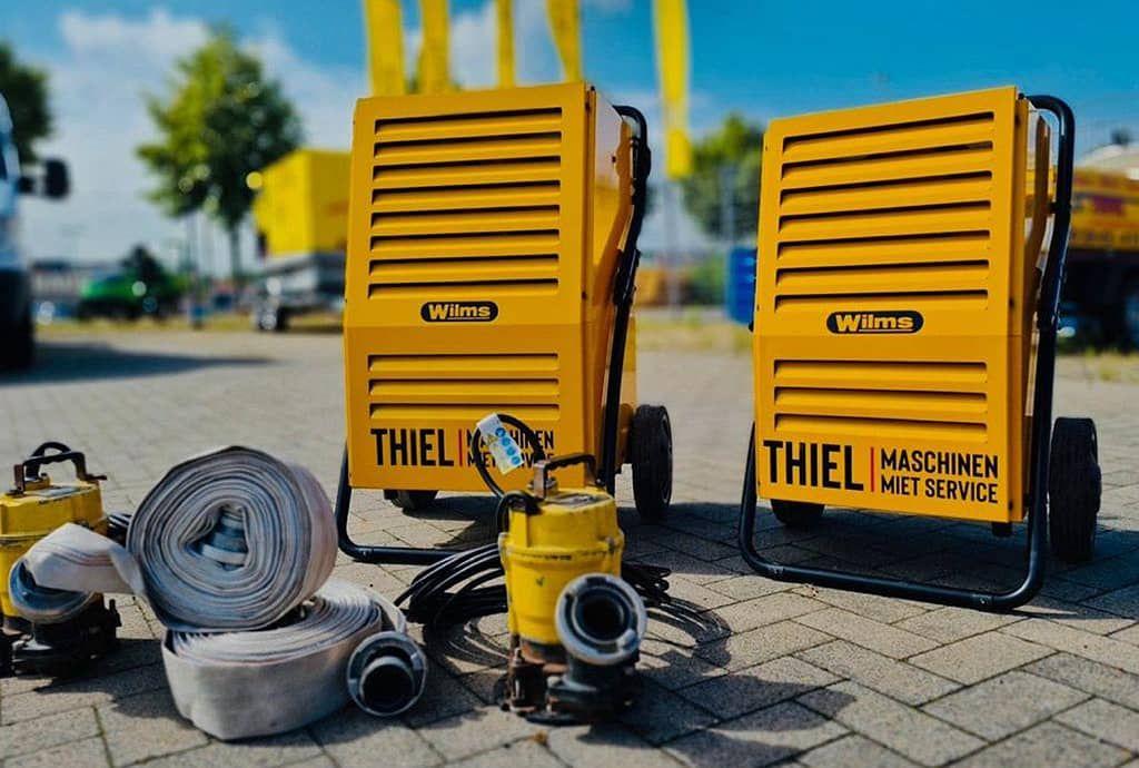 Zwei gelbe Wilms-Bautrockner der Firma Thiel Maschinenvermietung stehen auf gepflastertem Boden, daneben liegen zusammengerollte Wasserschläuche und gelbe Pumpentechnik für die Baustellenentwässerung.