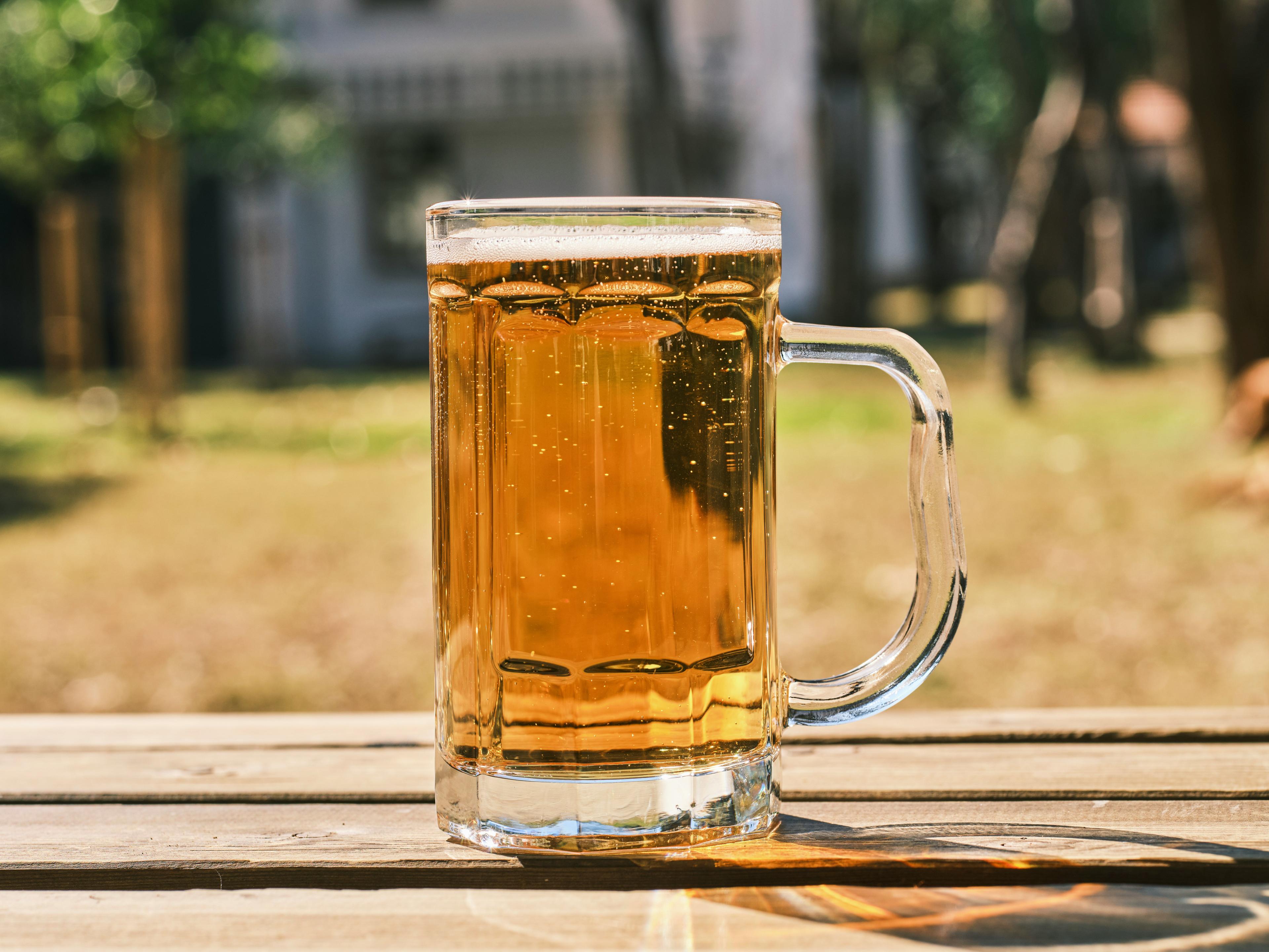 Goldenes Bierglas mit Henkel steht auf einem Holztisch im Freien, gefüllt mit hellem schäumendem Bier und Kohlensäurebläschen, vor unscharfem grünem Park- oder Gartenhintergrund bei Sonnenschein.