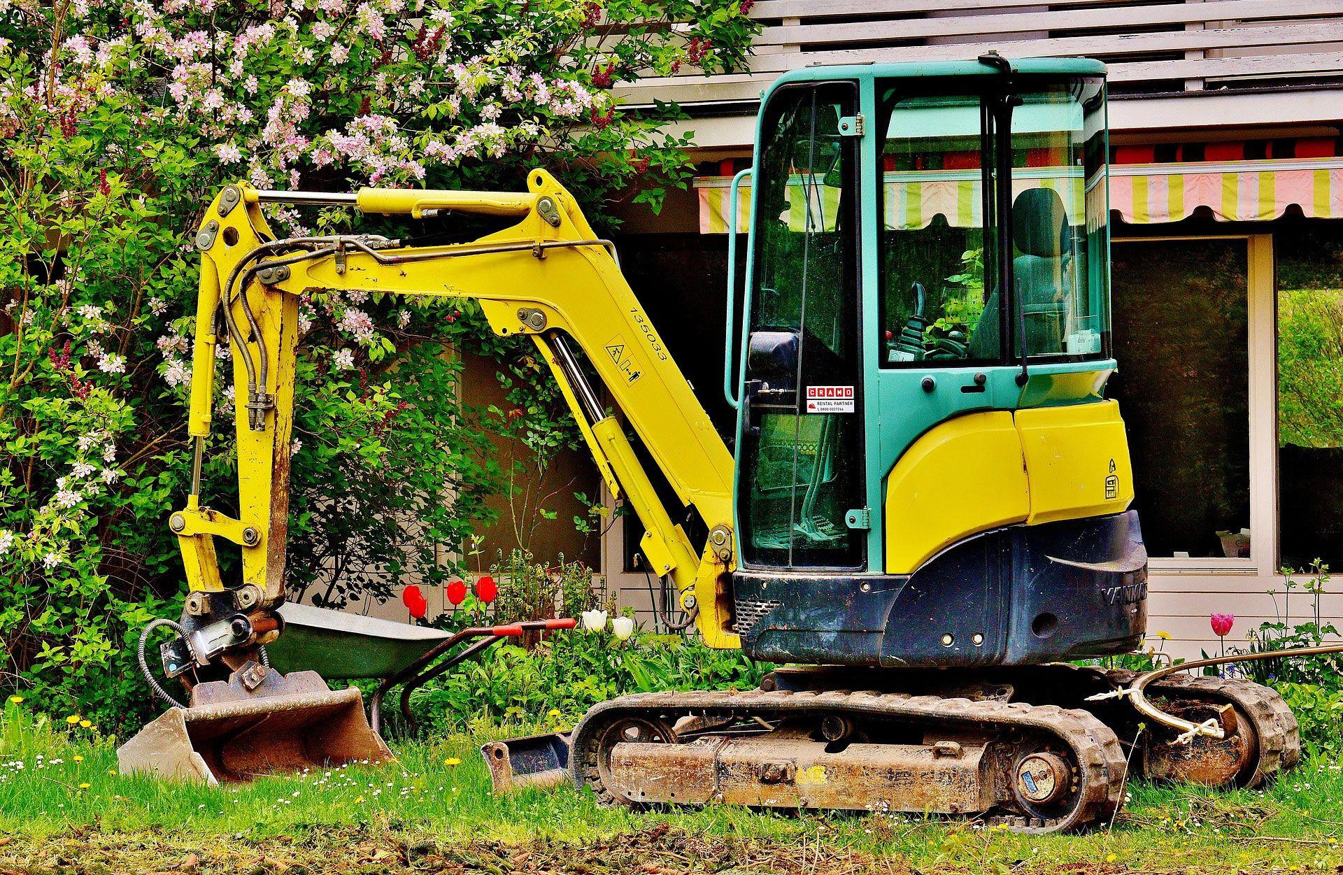 Kompakter gelb-grüner Minibagger steht in einem Garten vor einem Wohnhaus mit blühenden Sträuchern, Markise und gepflegtem Rasen, bereit für Gartenarbeiten oder kleinere Erdarbeiten im privaten Bereich.