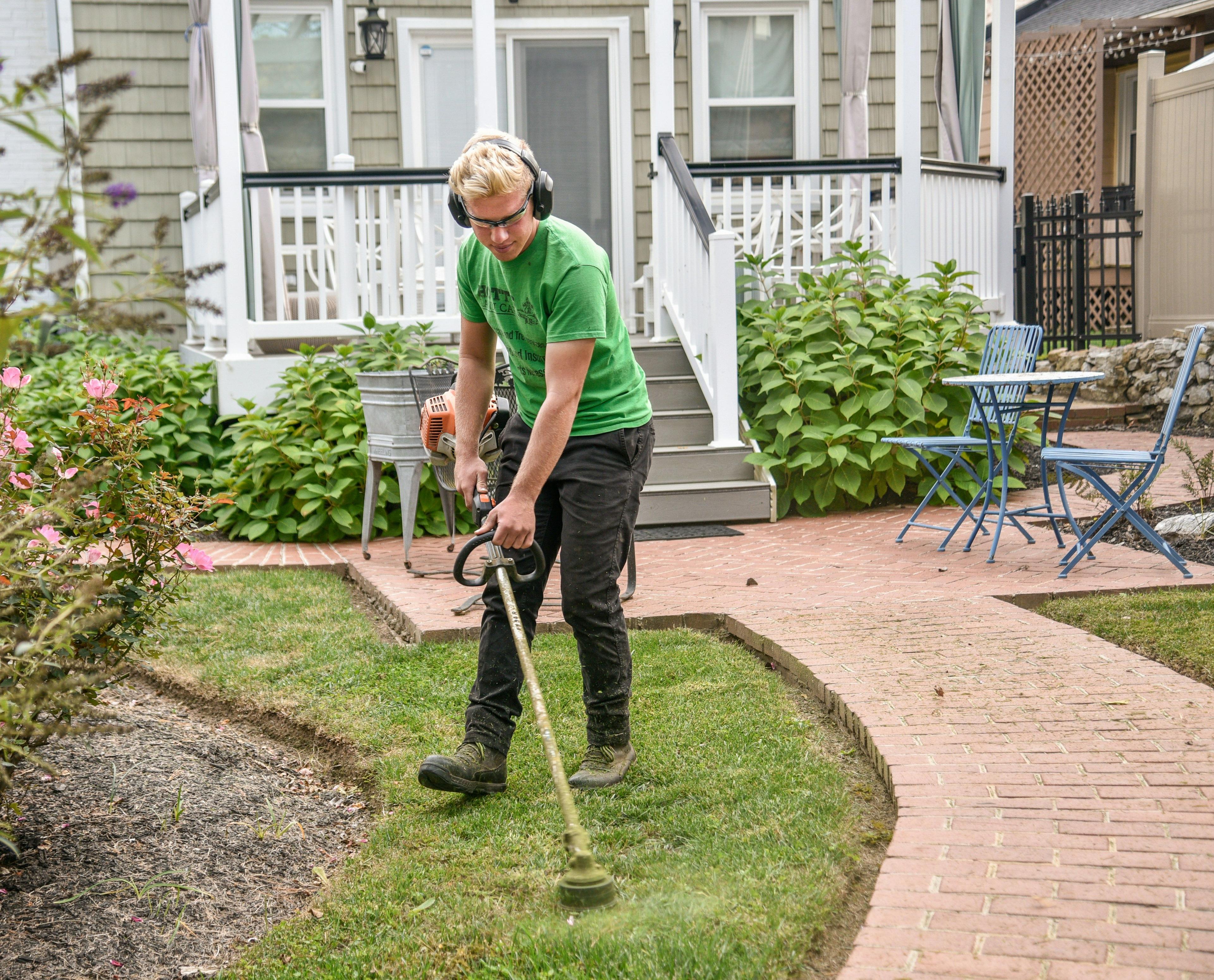 Mann in grünem T-Shirt und Gehörschutz trimmt mit einem Rasentrimmer präzise die Rasenkanten entlang eines gepflasterten Gartenwegs vor einem amerikanischen Holzhaus mit weißer Veranda, umgeben von gepflegten Beeten und blauen Gartenstühlen.