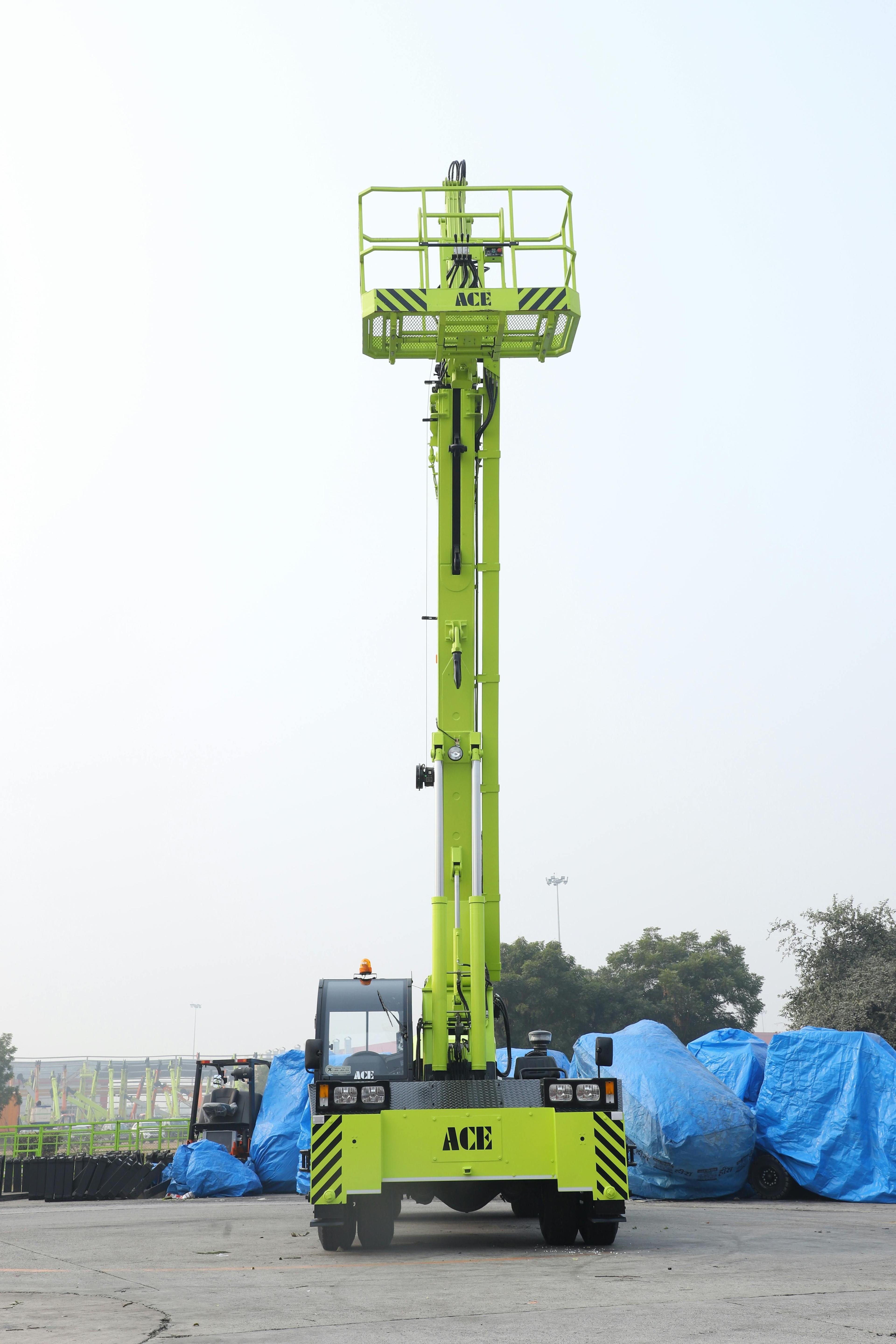 Grüne ACE-Teleskoparbeitsbühne mit hohem ausgefahrenem Mast und Arbeitskorb steht auf einem Betriebshof, im Hintergrund sind mit blauen Planen abgedeckte Maschinen und Bäume unter bewölktem Himmel zu sehen.