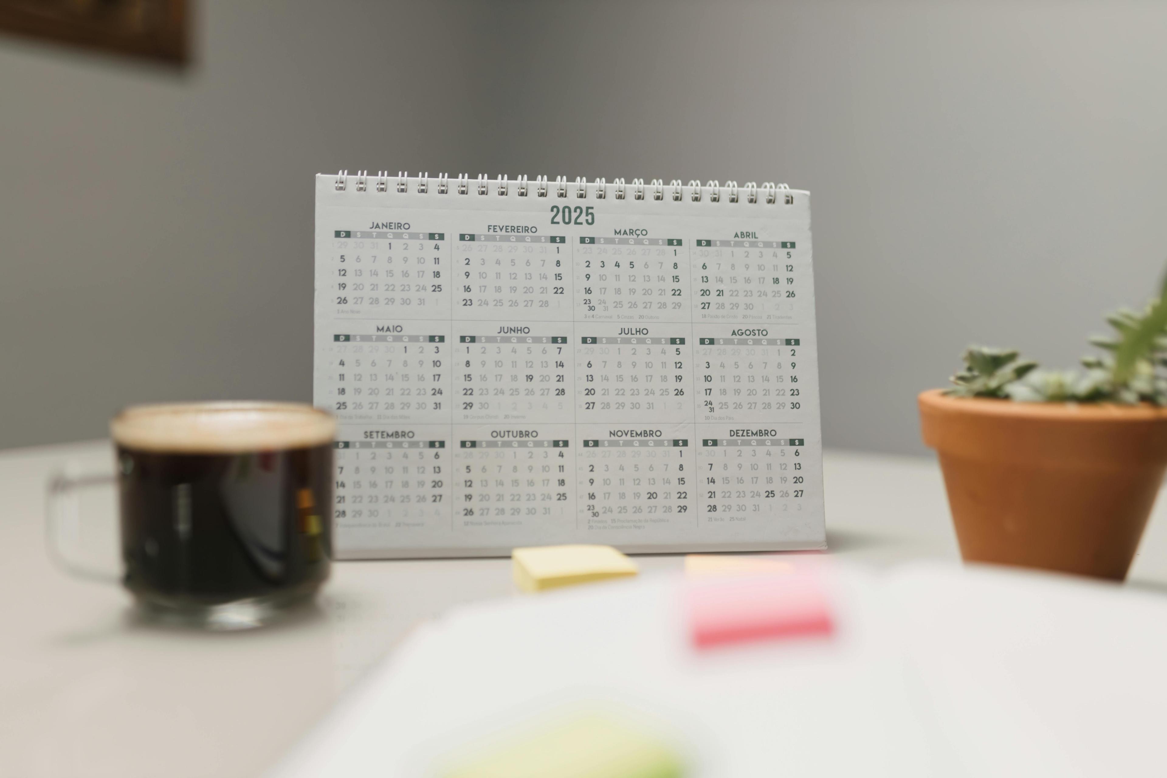 Aufstellkalender für 2025 mit portugiesischen Monatsnamen auf weißem Schreibtisch, daneben Kaffeetasse, Topfpflanze und bunte Haftnotizen.