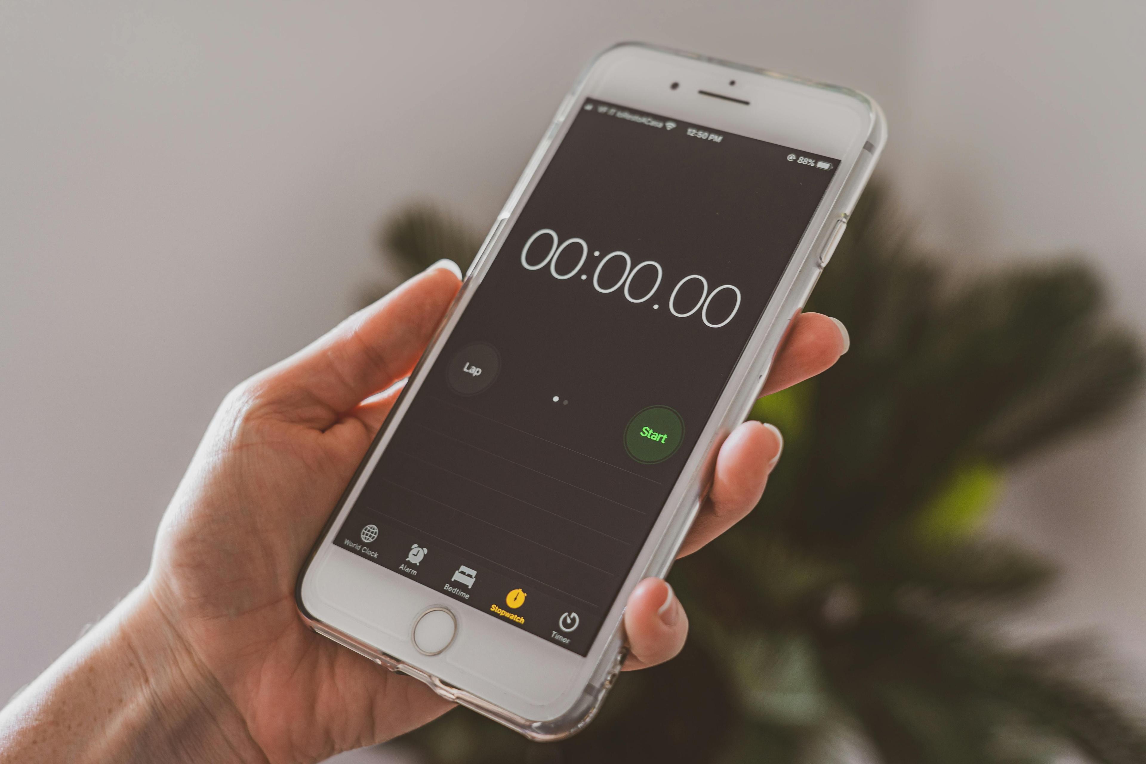 Hand hält weißes iPhone mit Stoppuhr-App auf dem Display, Zeit steht auf 00:00.00, grüner Start-Button sichtbar, unscharfer Innenraum-Hintergrund.