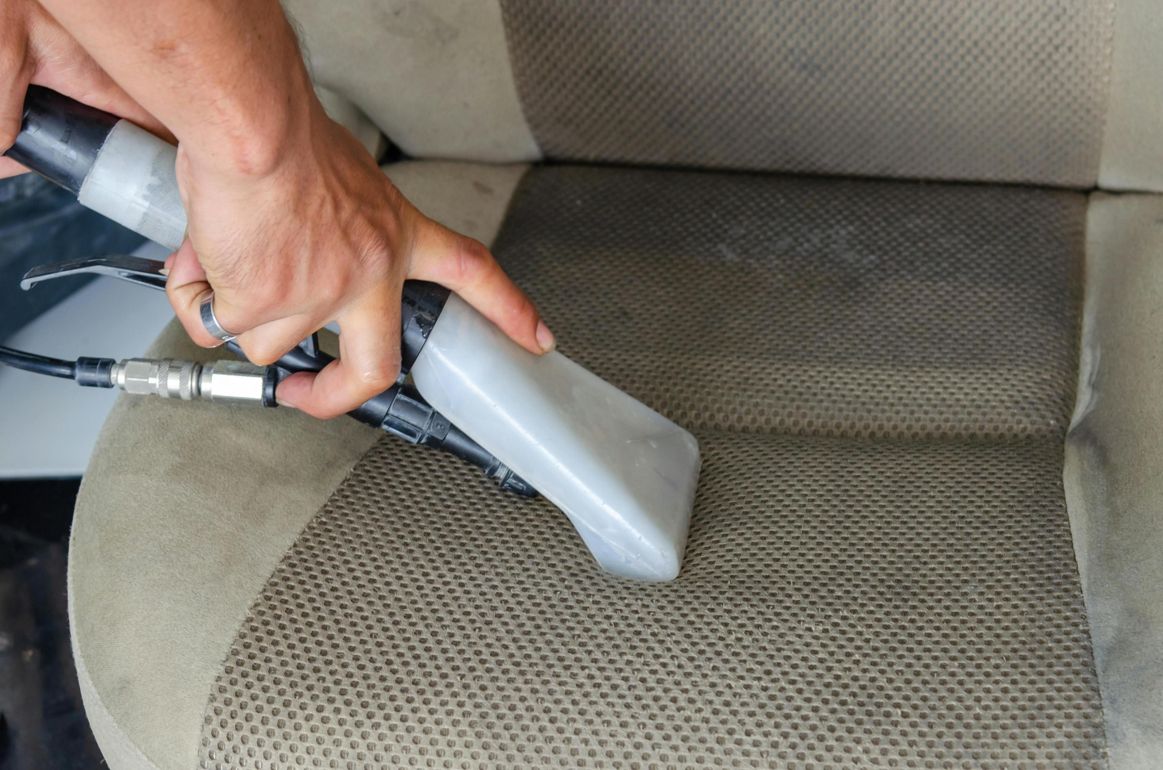 Hand hält Handstaubsauger mit transparentem Behälter und reinigt beige Autopolster mit perforiertem Muster.