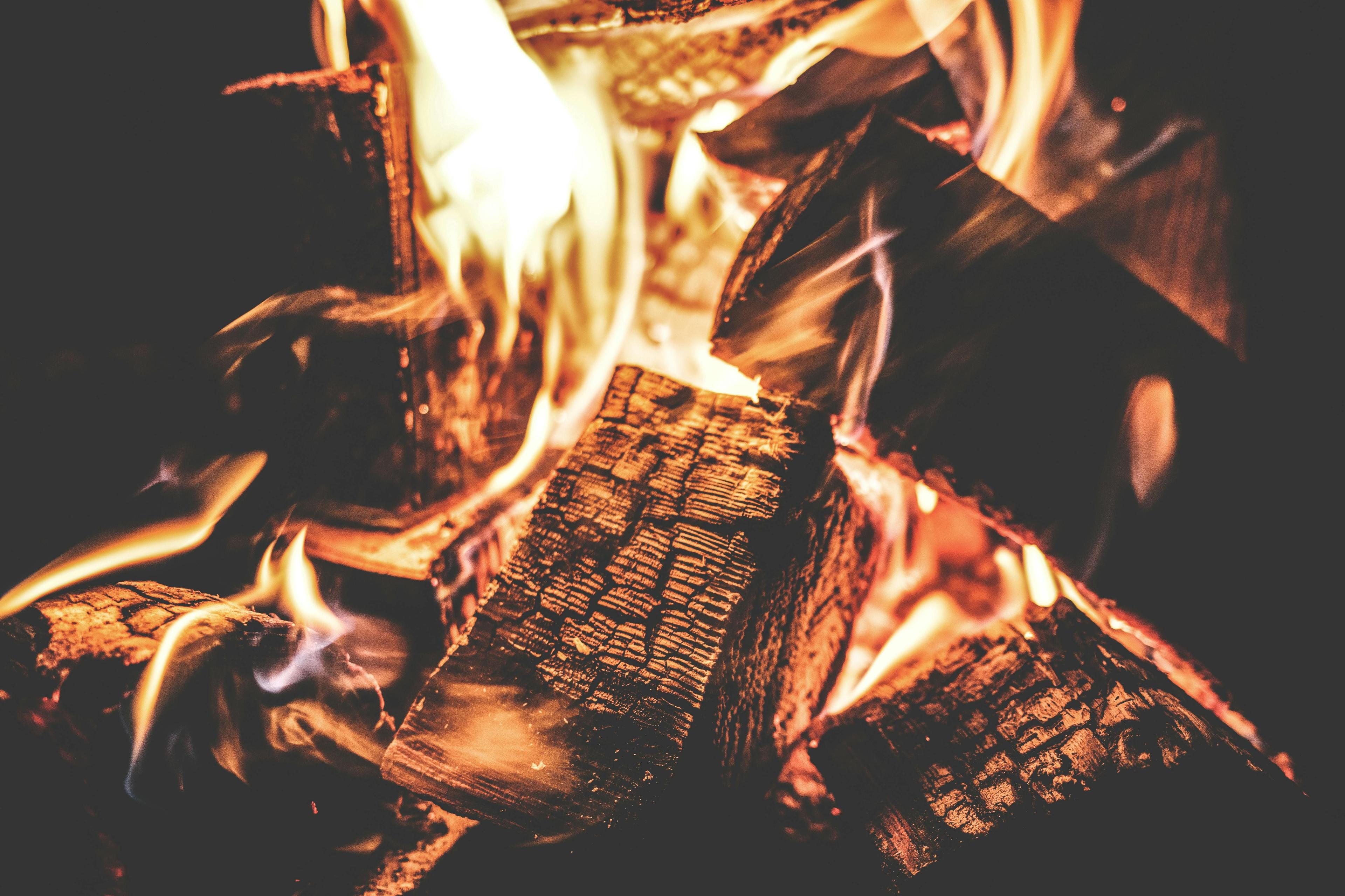 Loderndes Kaminfeuer mit hell brennenden Flammen, die um verkohlte Holzscheite züngeln, in warmen Orange- und Gelbtönen vor dunklem Hintergrund.