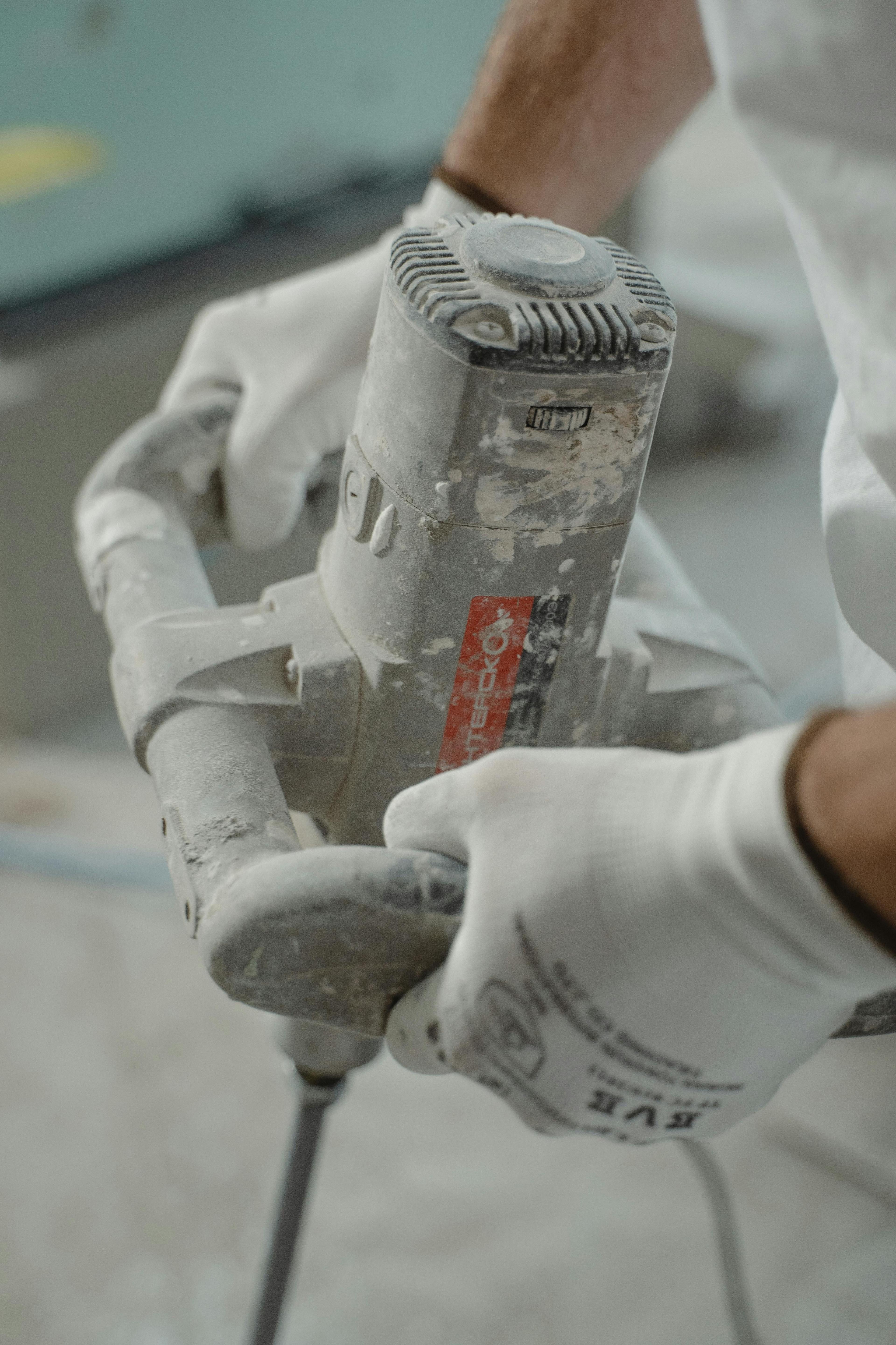Nahaufnahme von Handwerkerhänden in weißen Handschuhen, die einen staubbedeckten Winkelschleifer oder Bohrer bedienen, mit Staubwolken und Schleifpartikeln in der Luft während der Bearbeitung von Baumaterial..