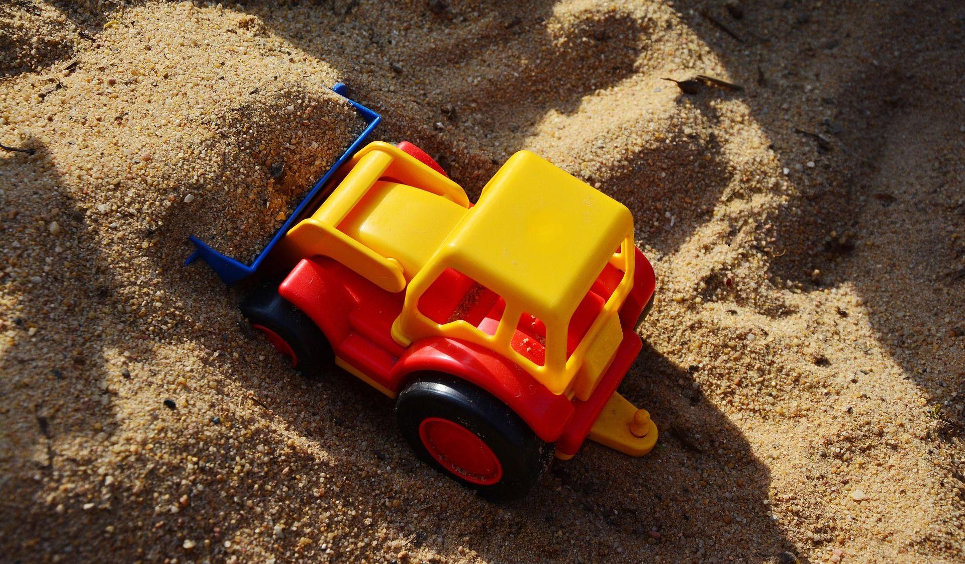 Buntes Kinderspielzeug-Kipplaster in rot-gelber Farbgebung mit schwarzen Rädern und blauer Schaufel liegt im Sand, umgeben von kleinen Sandhaufen auf einem Spielplatz oder im Sandkasten.