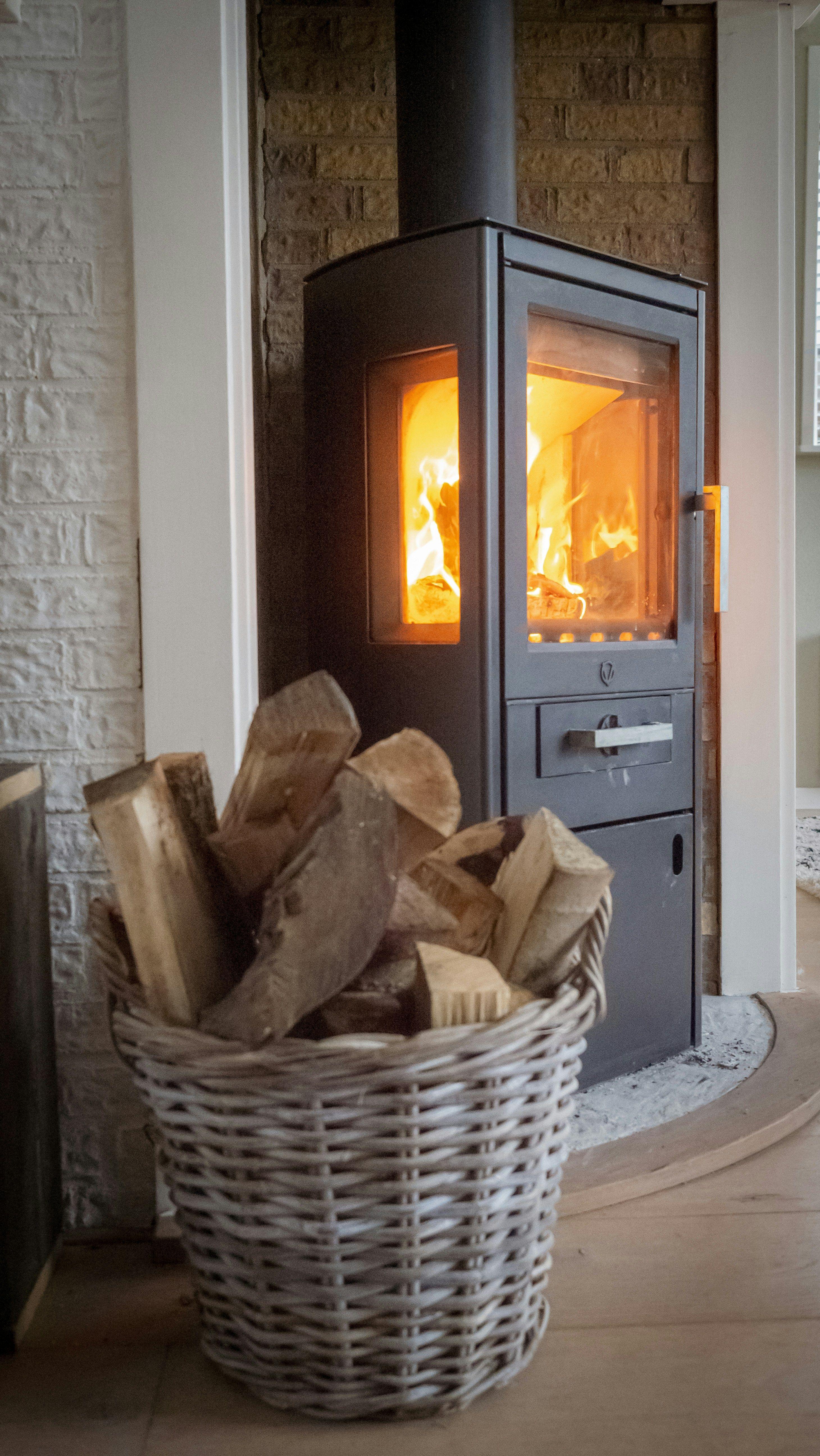 Moderner schwarzer Kaminofen mit brennendem Feuer hinter Glasscheibe, daneben steht ein geflochtener Weidenkorb voller Brennholz vor einer Natursteinwand in einem gemütlichen Wohnraum.