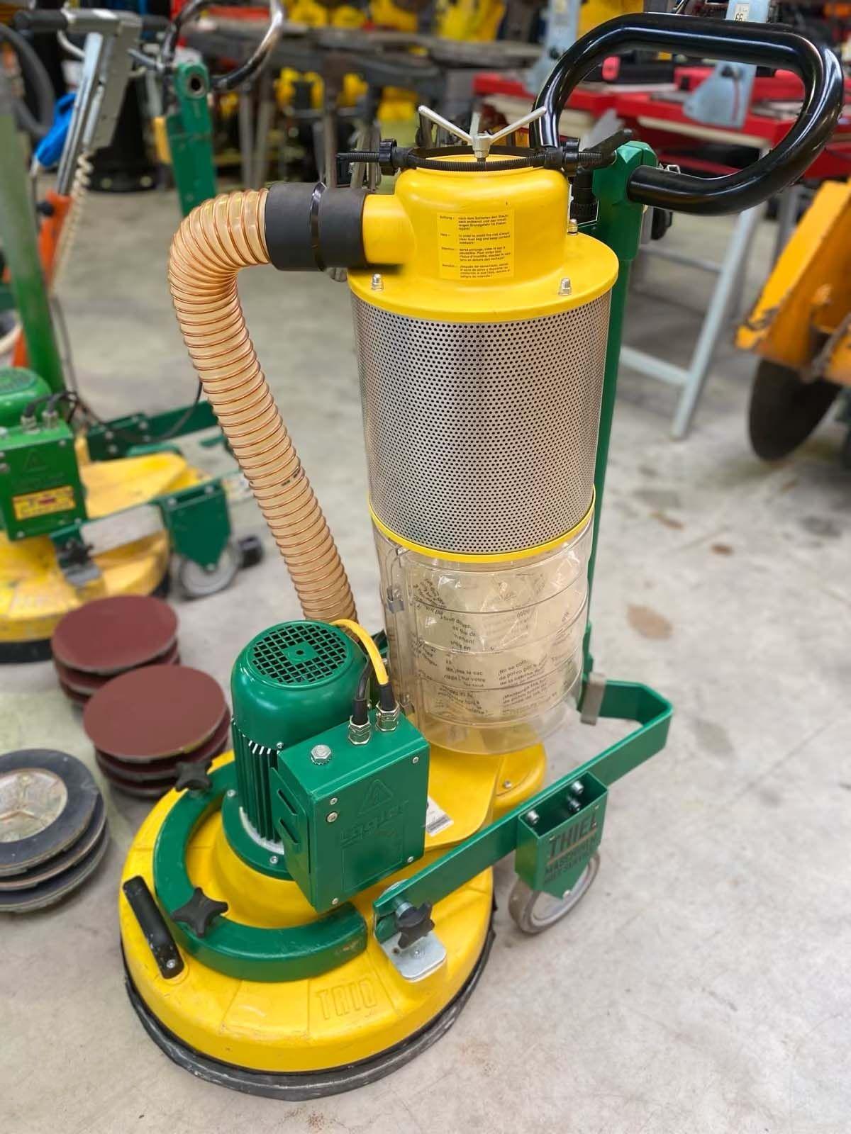 Gelb-grüne Bodenschleifmaschine mit Staubabsaugung, perforiertem Metallfilter, transparentem Auffangbehälter und Spiralschlauch in Werkstatthalle.