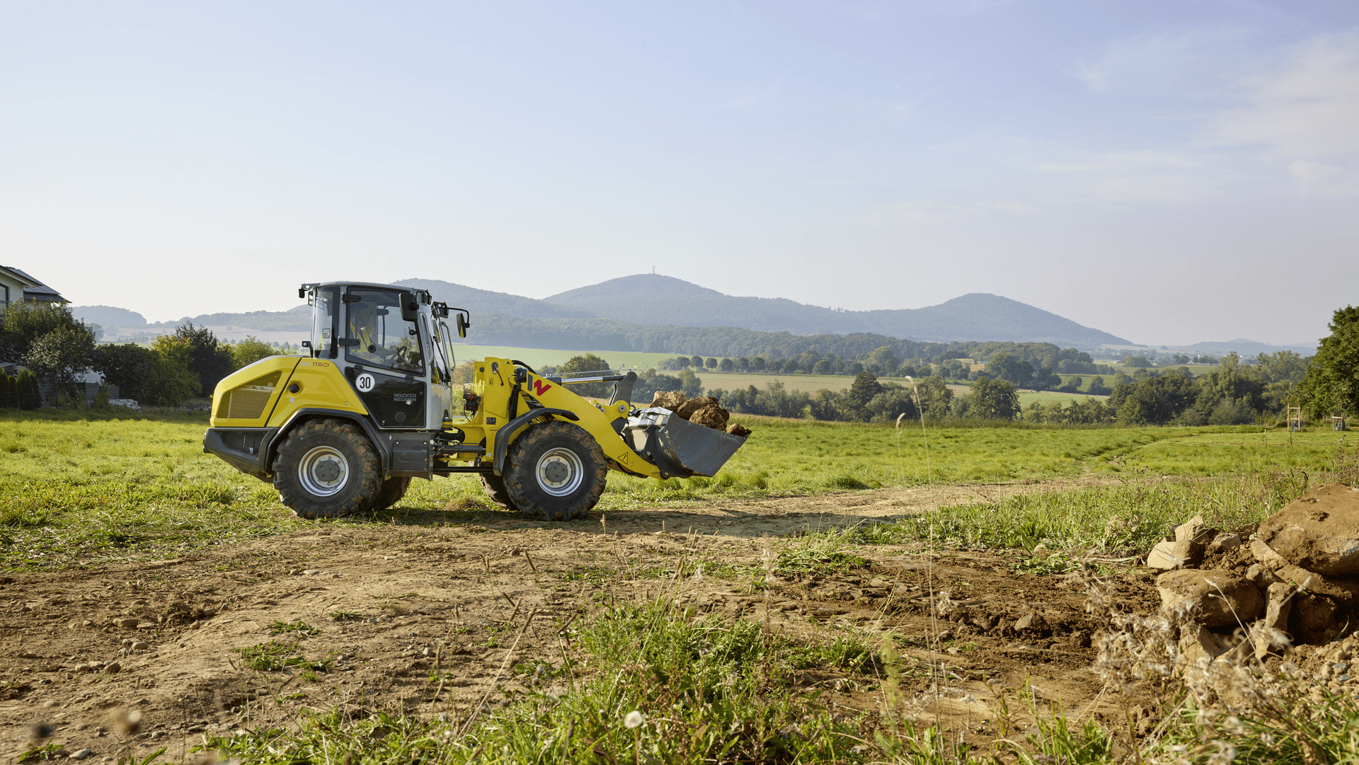 Gelber Kompaktlader mit Schaufel arbeitet auf einer ländlichen Baustelle oder einem Feld, transportiert Erdmaterial vor einer malerischen Landschaft mit grünen Hügeln, Wäldern und bewölktem Himmel im Hintergrund.