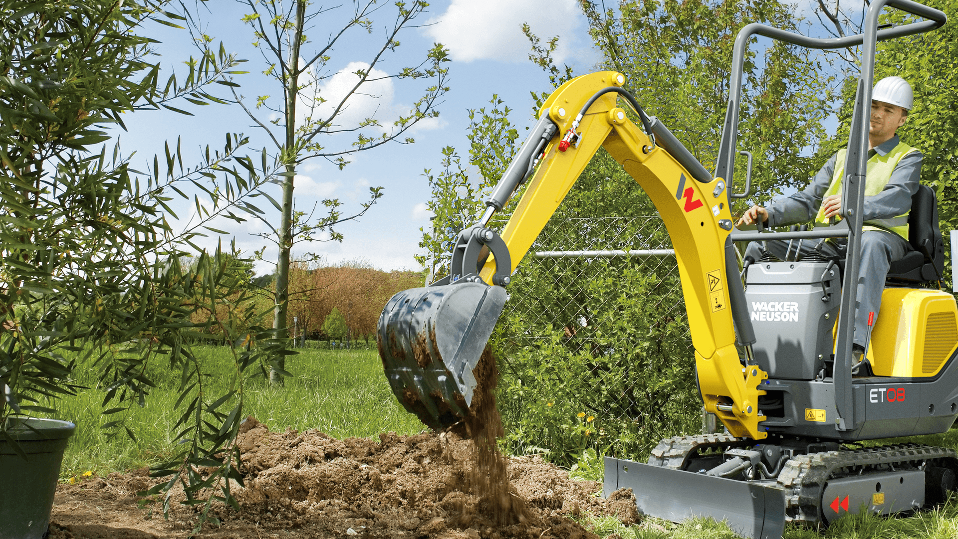 Arbeiter in Warnweste und Helm bedient gelben Wacker Neuson ET26 Minibagger beim Graben in grüner Gartenlandschaft mit jungen Bäumen, blauer Himmel im Hintergrund